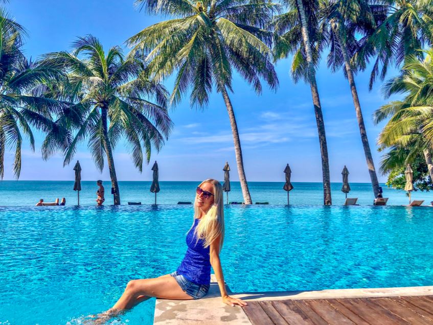 Frau sitzt vor Pool mit Palmen und Meer im Hintergrund