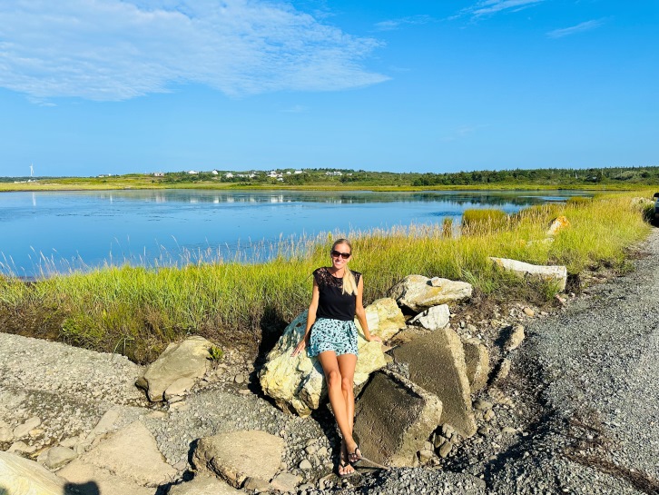 Cape Breton Titelbild, Frau sitzt auf Felsen, im Hintergrund grünes Gras und Wasser