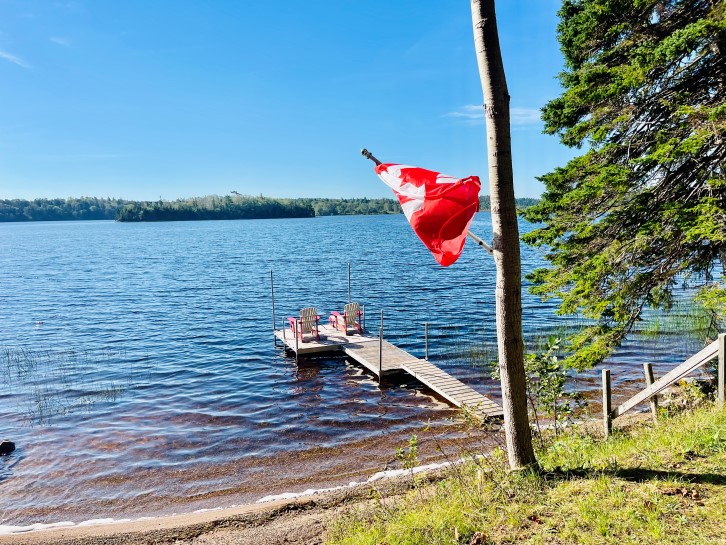 Kanadaflagge vor Holzsteg am See mit blauem Himmel und Sonnenschein auf Cape Breton